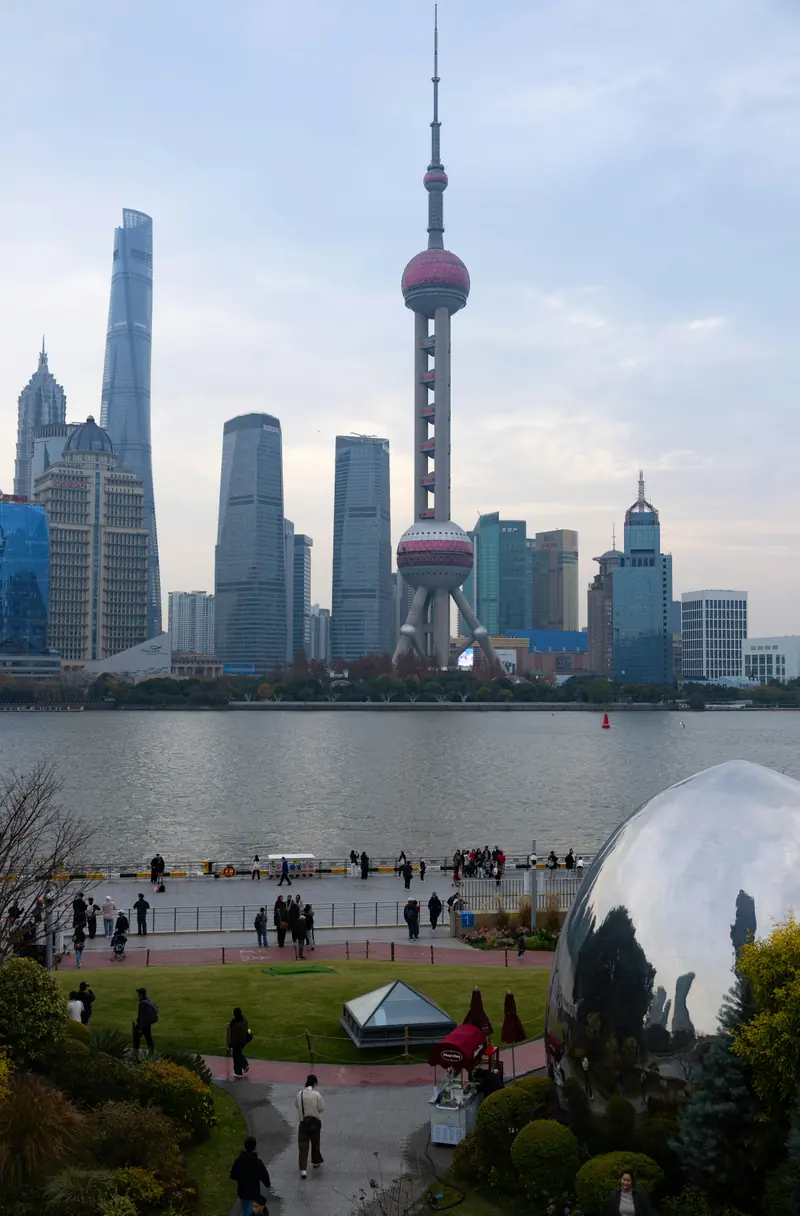 The Shanghai skyline across the Huangpu River, with the Oriental Pearl Tower and Lujiazui towers, and people walking along the riverside